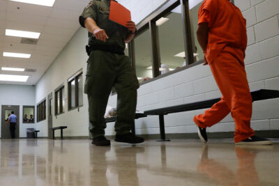 A prison guard walks a detainee with his hands behind his back to an intake area at an ICE Processing Center.